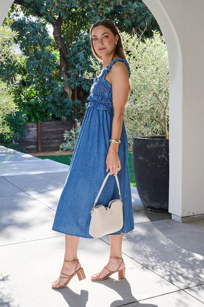 Woman in a Denim Ruffle Smocked Midi Dress holds a white purse outdoors under an archway in the sunlight.