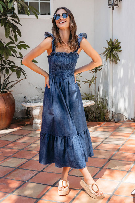 Woman outdoors in a Denim Ruffle Tiered Midi Dress, sandals and sunglasses, smiling on a tiled patio.