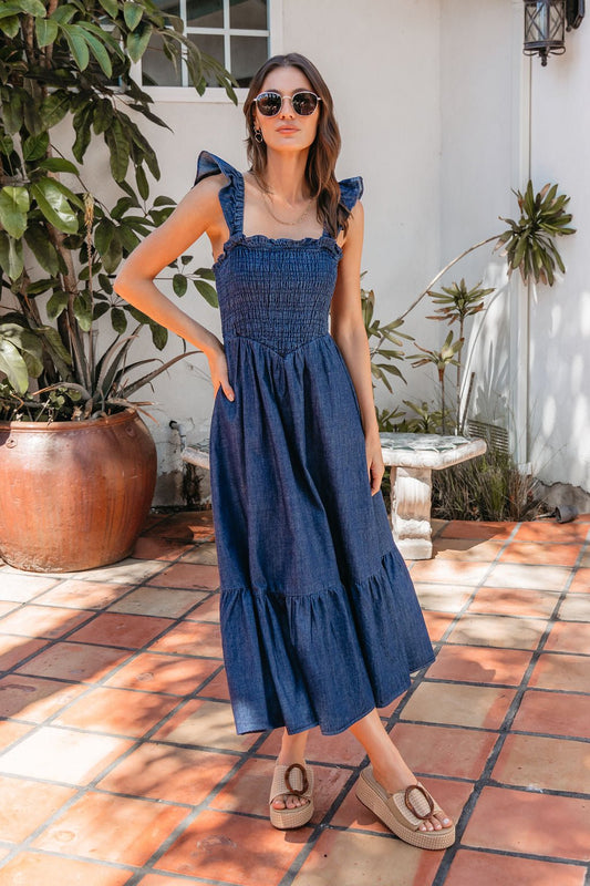 Woman outdoors in a Denim Ruffle Tiered Midi Dress, sunglasses, platform sandals, surrounded by plants and terracotta tiles.