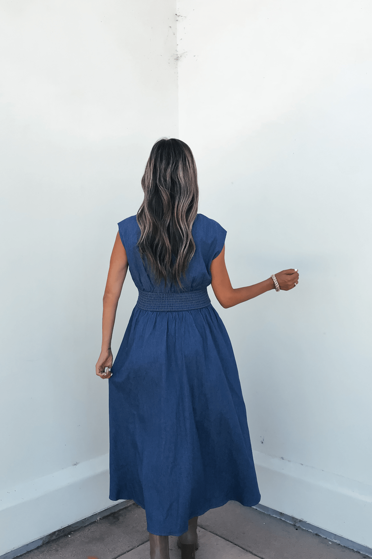 Woman with long wavy hair faces a white wall, wearing the Denim Smocked Zip Up Midi Dress - FINAL SALE, back to camera.