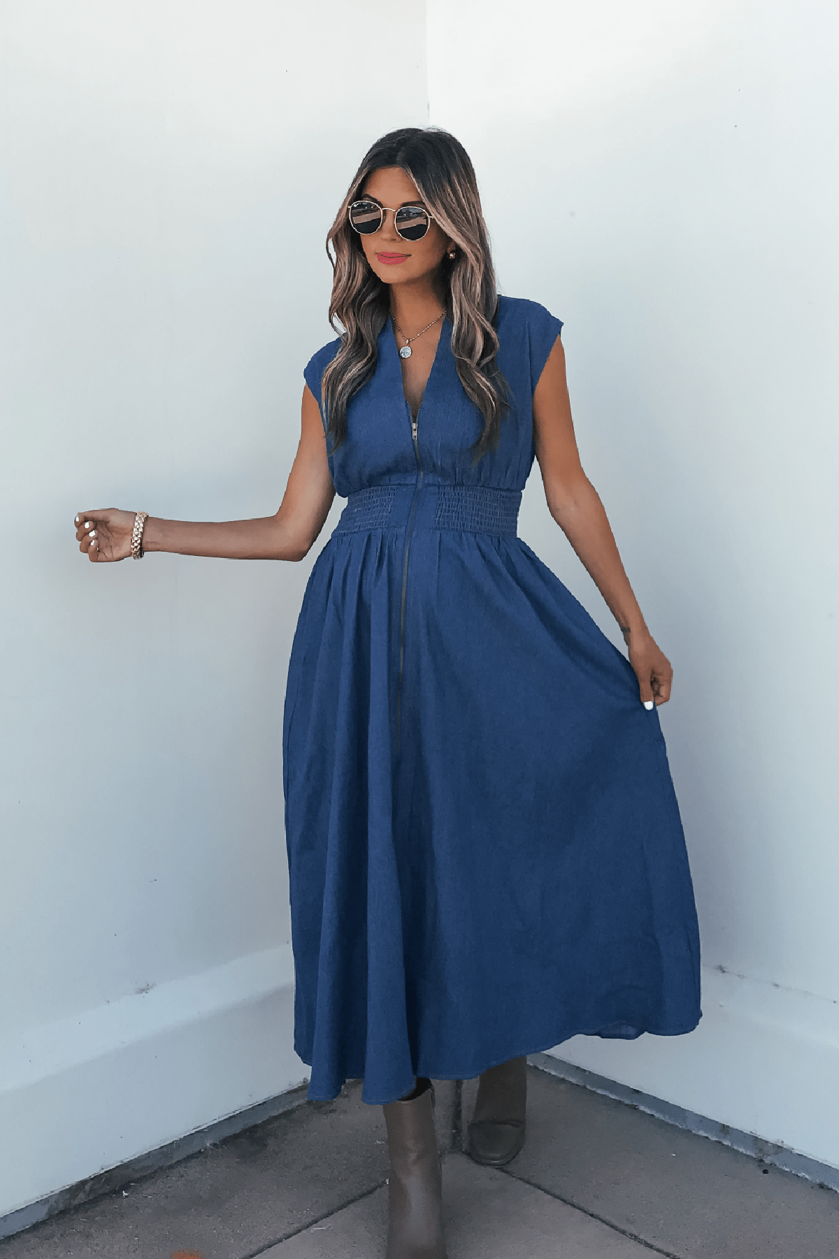 Woman in a Denim Smocked Zip Up Midi Dress poses stylishly with sunglasses, long wavy hair, and taupe boots against a white wall.