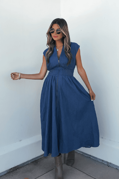 Woman in a Denim Smocked Zip Up Midi Dress poses stylishly with sunglasses, long wavy hair, and taupe boots against a white wall.