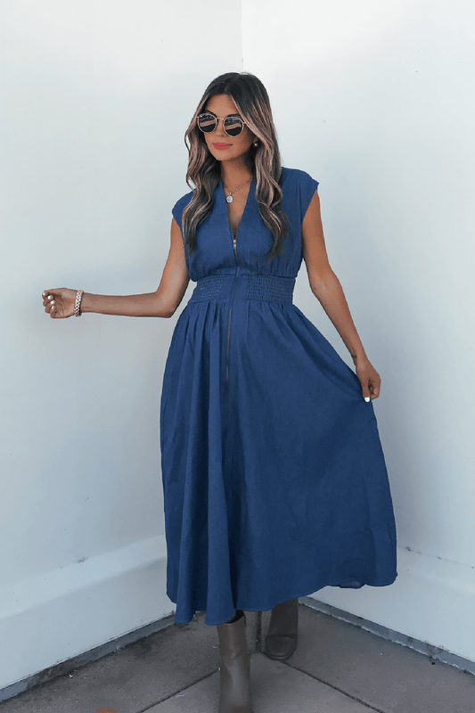 Woman in the Denim Smocked Zip Up Midi Dress stands against a white wall, holding her skirt, with sunglasses and brown boots.