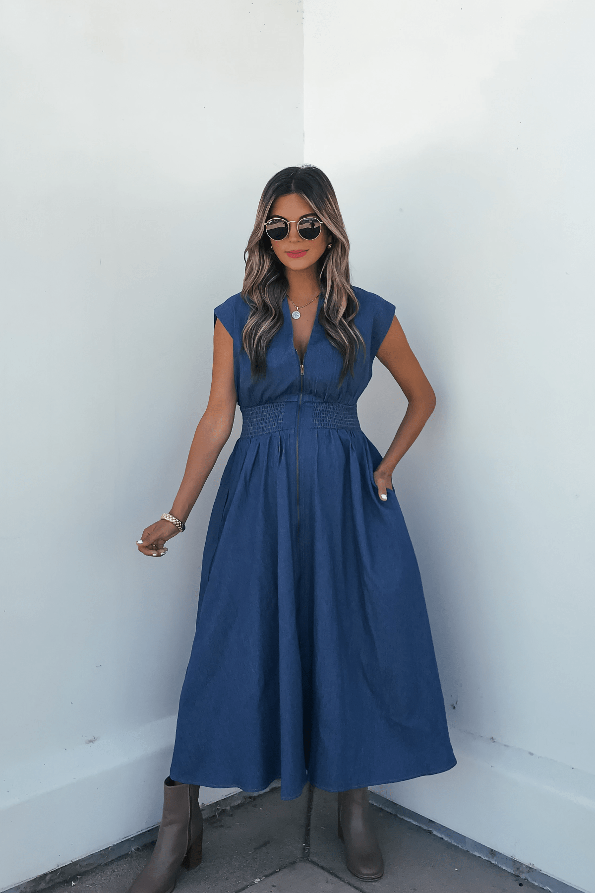 Woman in Denim Smocked Zip Up Midi Dress - FINAL SALE, sunglasses, brown boots, standing by a white wall with one hand in her pocket.