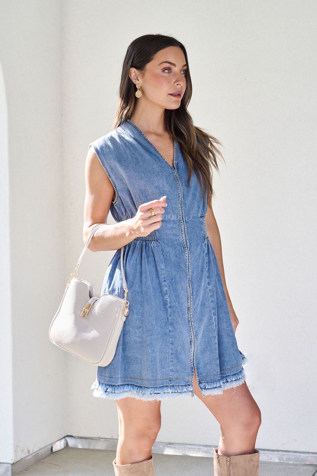Woman wearing the Denim Smocked Zip Up Mini Dress, holding a white handbag, standing indoors by a light wall.