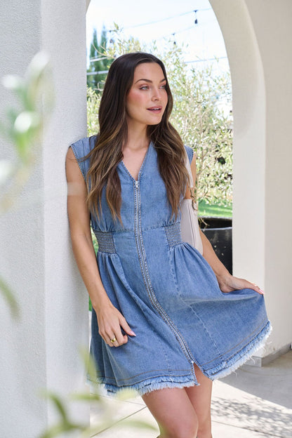 Woman wearing the Denim Smocked Zip Up Mini Dress stands by a white archway, holding the skirt with a relaxed side glance.