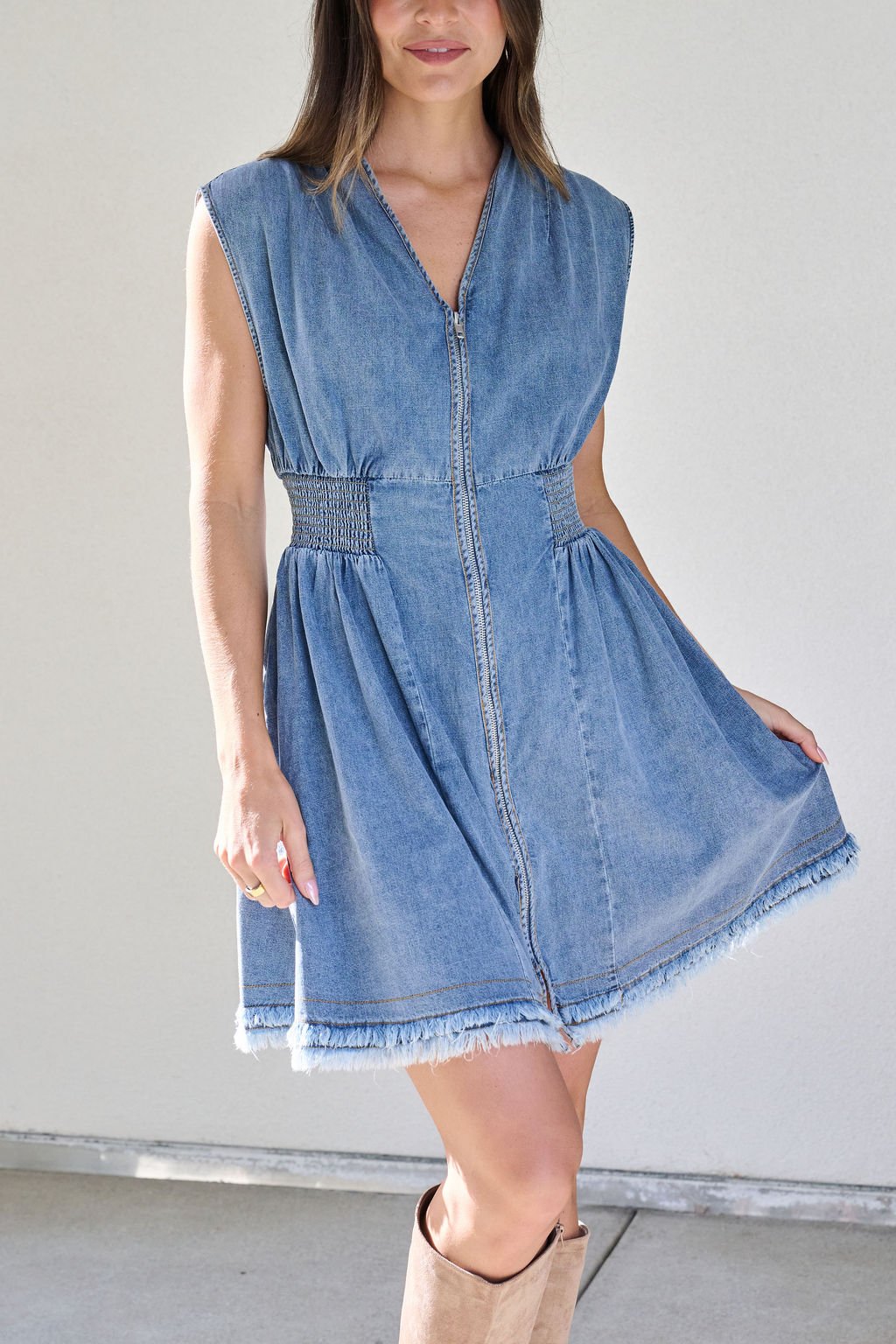 A woman in the Denim Smocked Zip Up Mini Dress with a frayed hem, smocked waist, zip front, and tan knee-high boots.