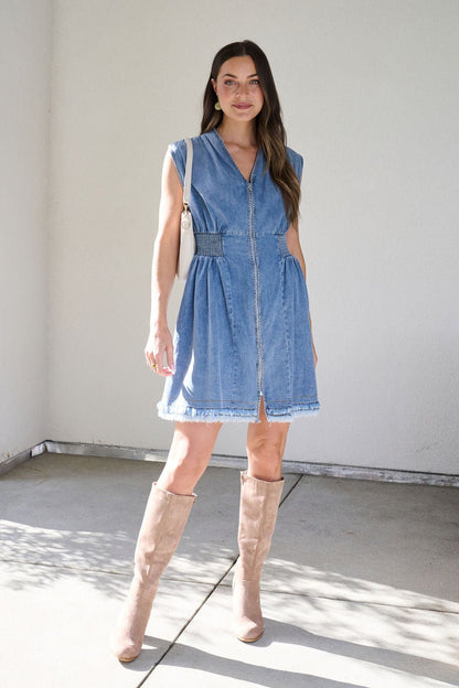 A woman wears the Denim Smocked Zip Up Mini Dress with tan knee-high boots, smiling at the camera against a white wall.