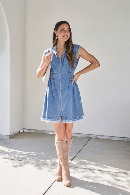 Woman in a Denim Smocked Zip Up Mini Dress and tan knee-high boots smiles, holding a white bag against a light wall.