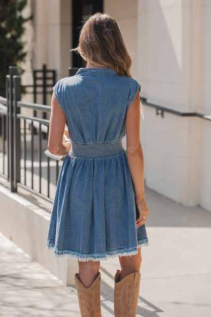 Woman outdoors in the Denim Smocked Zip Up Mini Dress with a frayed hem and tan cowboy boots, facing away.