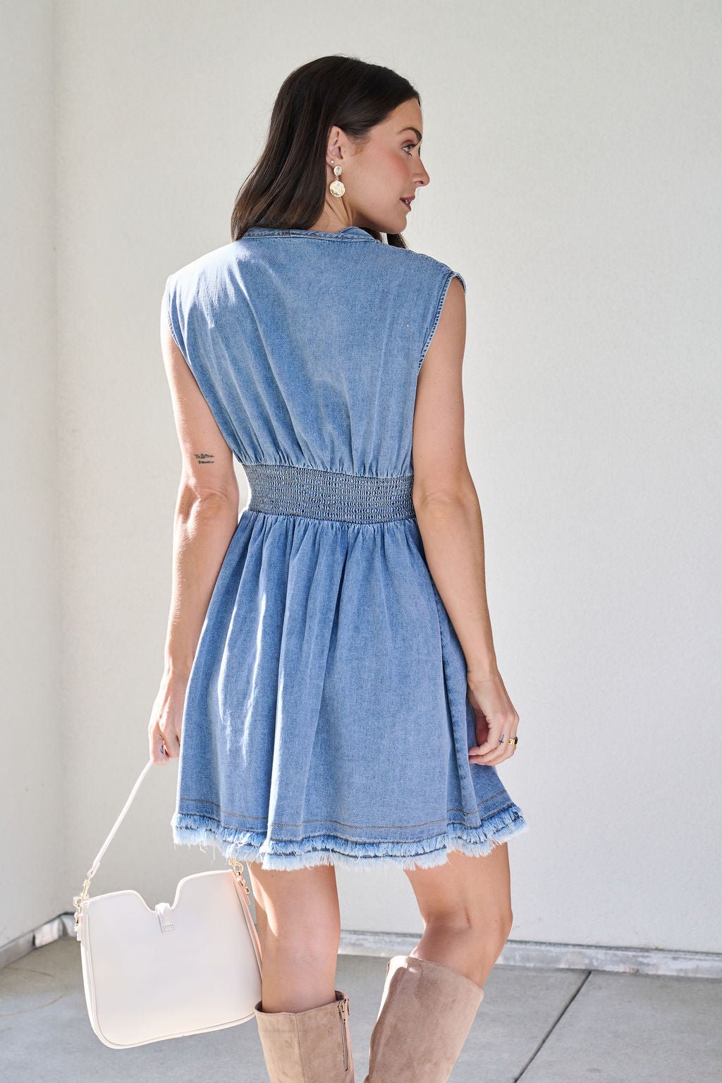 A woman in a Denim Smocked Zip Up Mini Dress holds a white purse, facing away in beige knee-high boots.