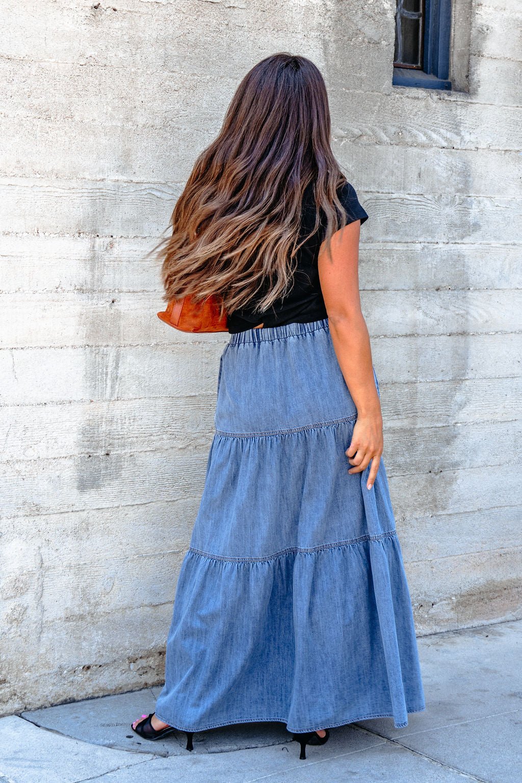 A woman in a black top and Denim Wash Tiered Maxi Skirt exudes casual chic while standing by a concrete wall, facing away.