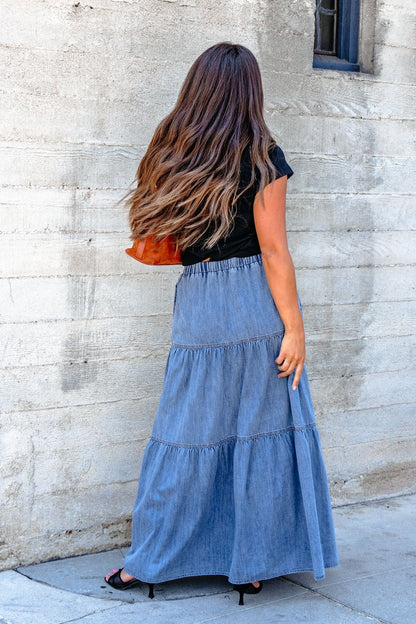 A woman in a black top and Denim Wash Tiered Maxi Skirt exudes casual chic while standing by a concrete wall, facing away.