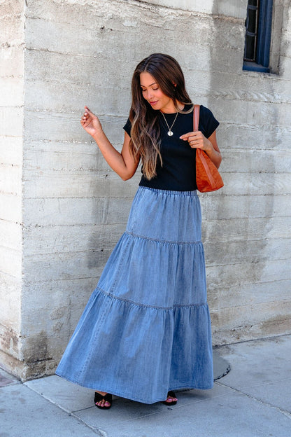 Woman in a black t-shirt and Denim Wash Tiered Maxi Skirt stands against a concrete wall, holding an orange bag for a casual chic look.
