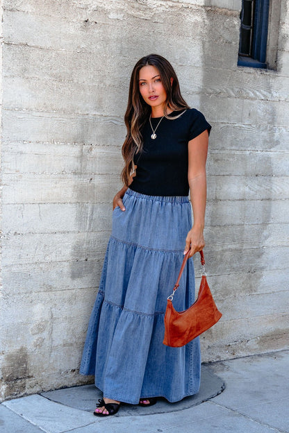 Woman with long hair in a black top wears the Denim Wash Tiered Maxi Skirt and holds an orange handbag by a concrete wall.