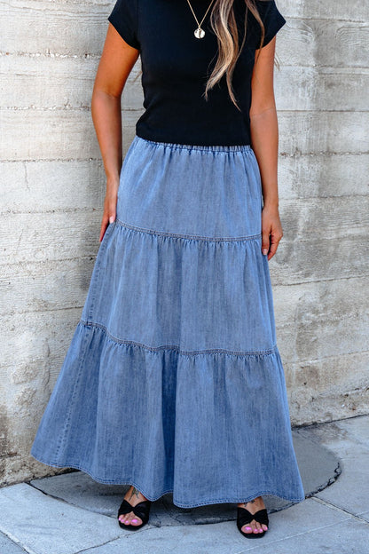A woman in a black top and Denim Wash Tiered Maxi Skirt stands against a concrete wall, hands on her casual chic skirt.
