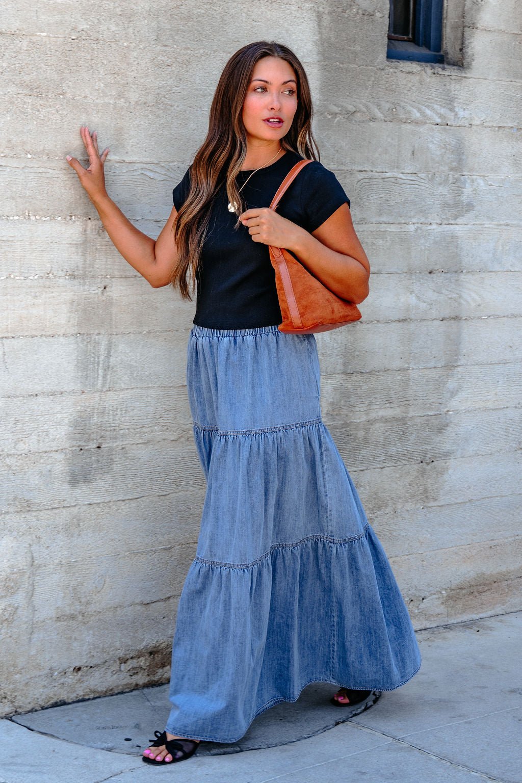 Woman in a black top and the Denim Wash Tiered Maxi Skirt stands by a concrete wall, carrying a brown bag with casual chic style.
