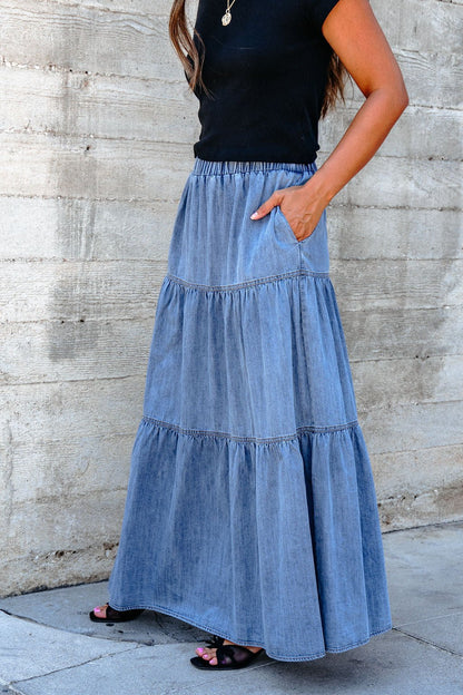 Woman in Denim Wash Tiered Maxi Skirt and black top exudes effortless casual chic against a concrete wall.