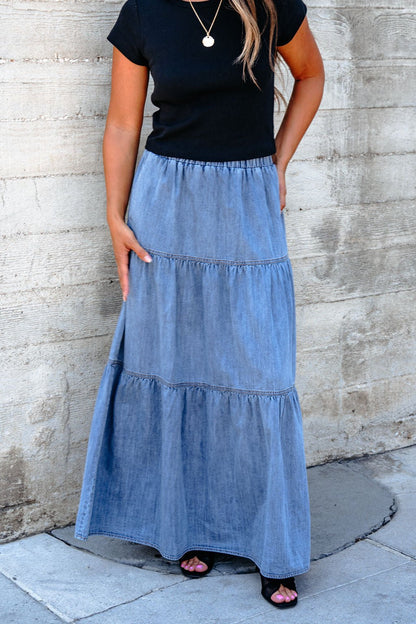 A woman in a black top and the Denim Wash Tiered Maxi Skirt exudes casual chic against a concrete wall.
