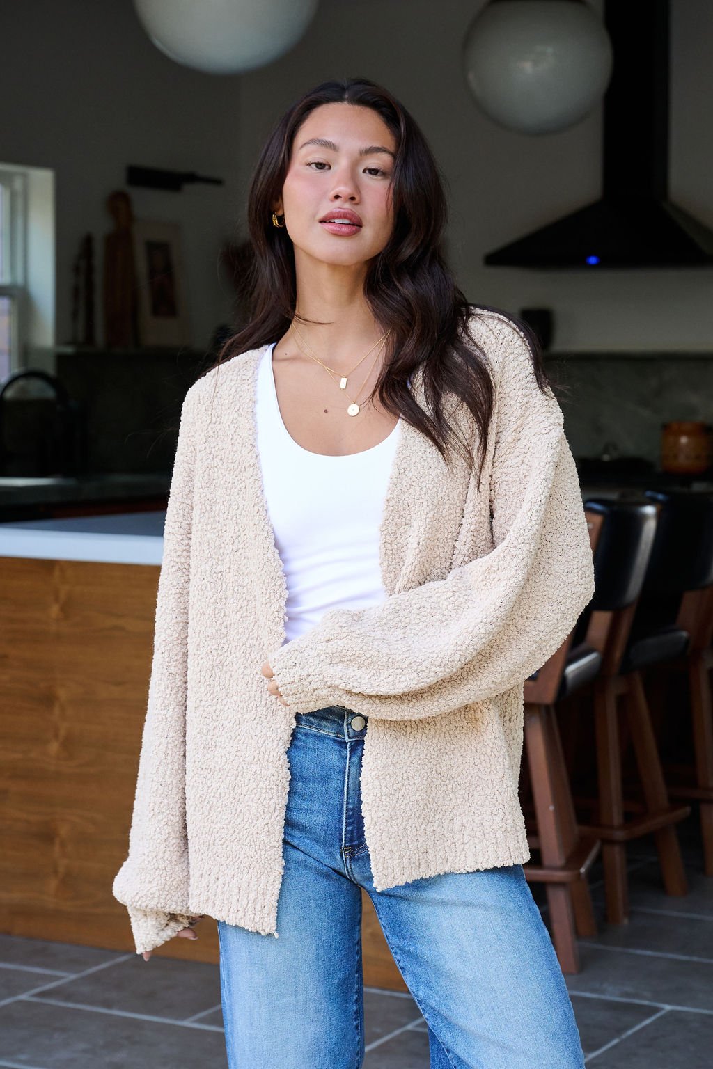 A woman wears the Desert Rose Popcorn Textured Open Front Cardigan with a white top and blue jeans, standing confidently in a modern kitchen.