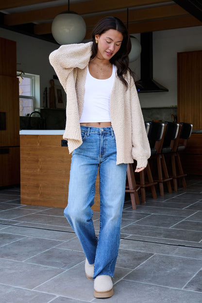 A woman in a Desert Rose Popcorn Textured Open Front Cardigan, white top, and blue jeans stands in a modern kitchen.