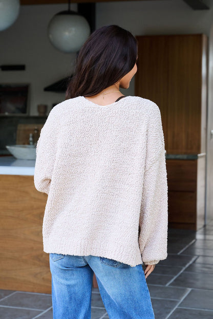 A woman with long dark hair, in blue jeans and a Desert Rose Popcorn Textured Open Front Cardigan, stands indoors with her back to the camera.