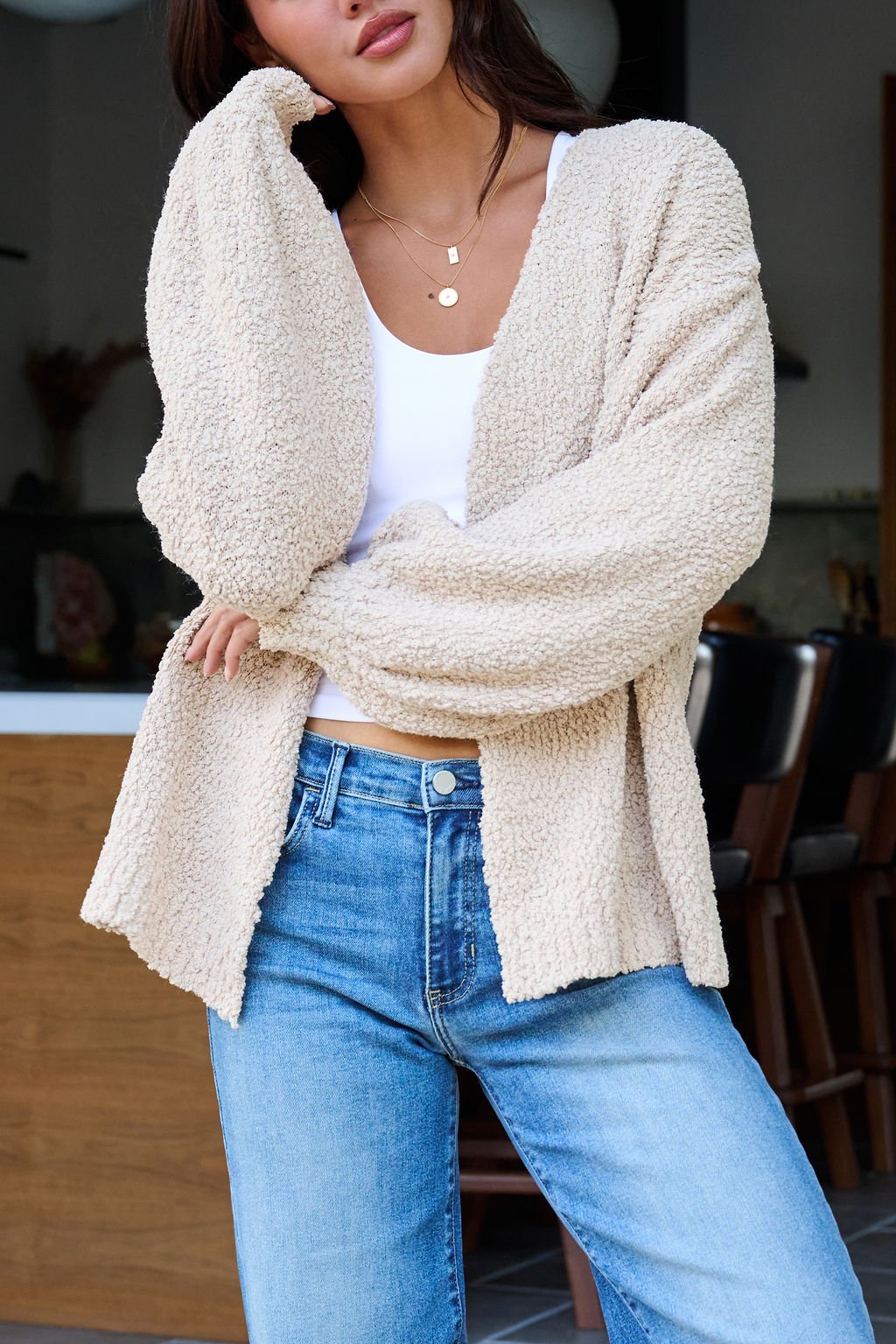 Woman in a Desert Rose Popcorn Textured Open Front Cardigan over a white top and blue jeans, standing indoors, hand to face.