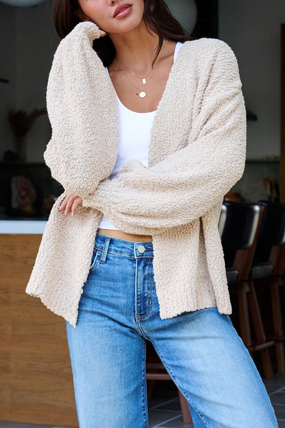 Woman in a Desert Rose Popcorn Textured Open Front Cardigan over a white top and blue jeans, standing indoors, hand to face.