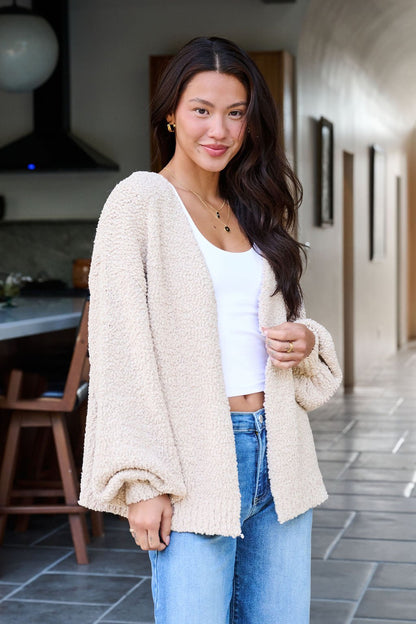 A woman in a Desert Rose Popcorn Textured Open Front Cardigan, white top, and blue jeans smiles in a modern kitchen.