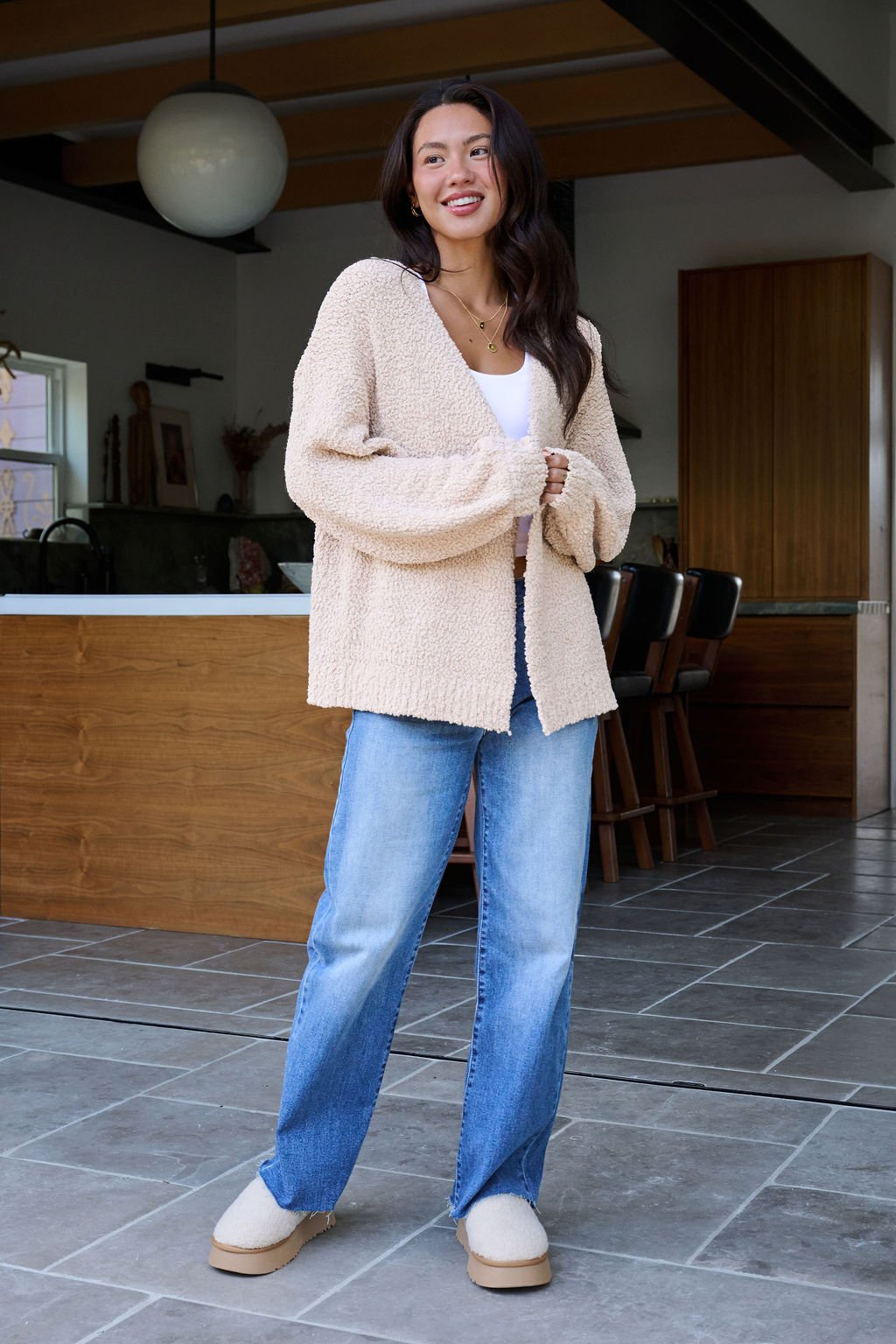 A woman in a Desert Rose Popcorn Textured Open Front Cardigan, white top, and blue jeans smiles in a modern kitchen.