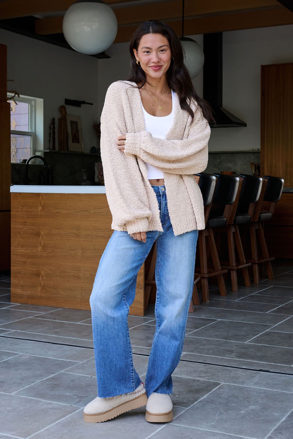 A woman in a Desert Rose Popcorn Textured Open Front Cardigan, white top, and blue jeans smiles in a modern kitchen.