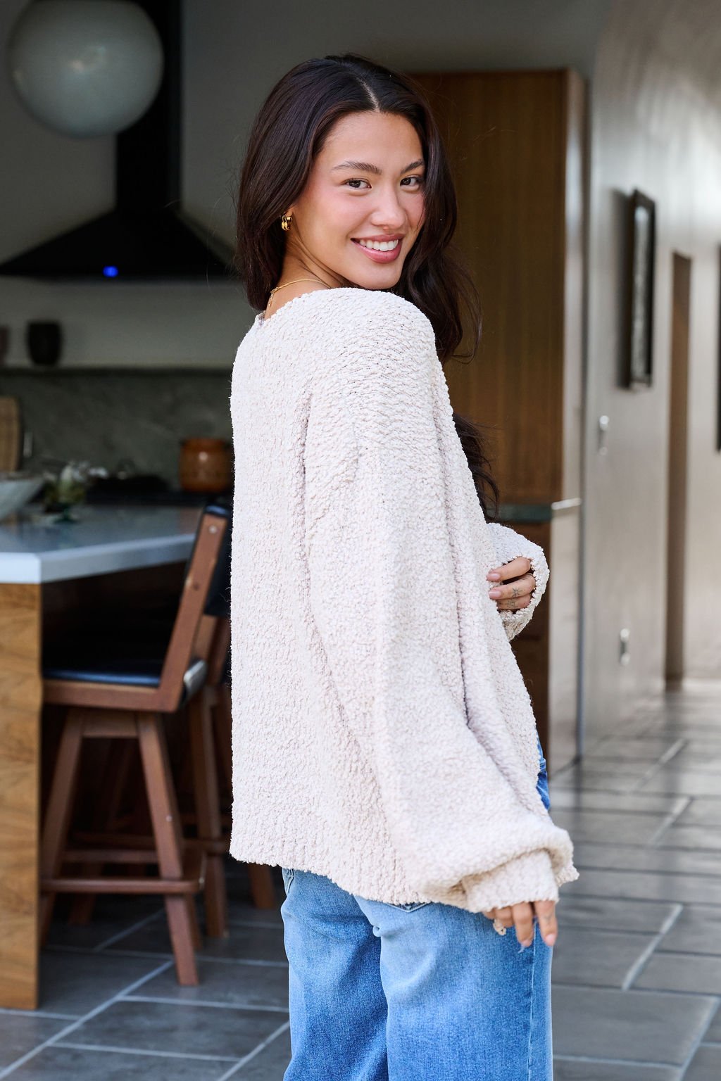 A woman in a Desert Rose Popcorn Textured Open Front Cardigan and jeans smiles while standing in a modern kitchen.