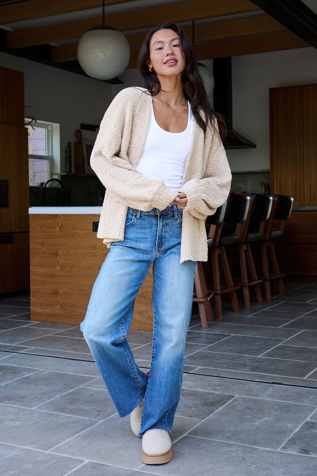 A woman in a Desert Rose Popcorn Textured Open Front Cardigan, white tank, blue jeans, and slippers stands in a modern kitchen.