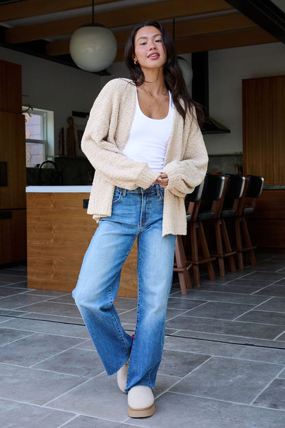 A woman in a Desert Rose Popcorn Textured Open Front Cardigan, white tank, blue jeans, and slippers stands in a modern kitchen.