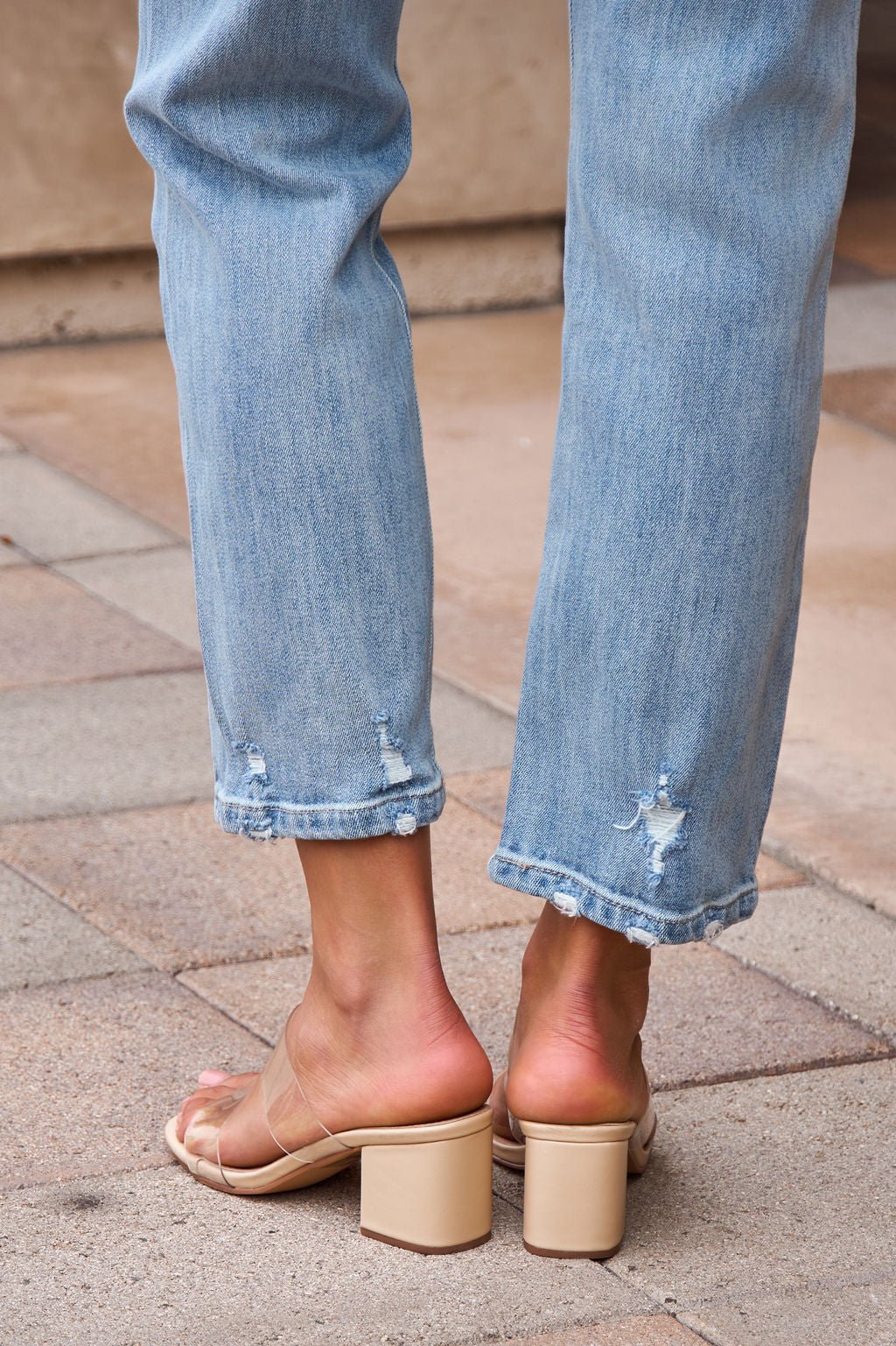 Someone wears Distressed Slim Straight Leg Jeans - Medium Wash and beige heeled sandals while standing on stone pavement.