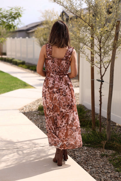 A woman wears the Drifter Brown Floral Print Tiered Maxi Dress, walking on a sidewalk beside a white fence and small trees.
