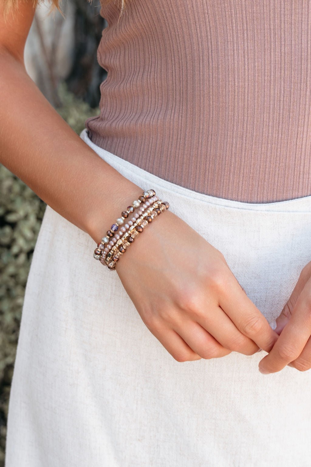 A woman in a ribbed mauve top and light skirt models the Driftwood Dreams Beaded Layered Bracelets, ideal for summer style.
