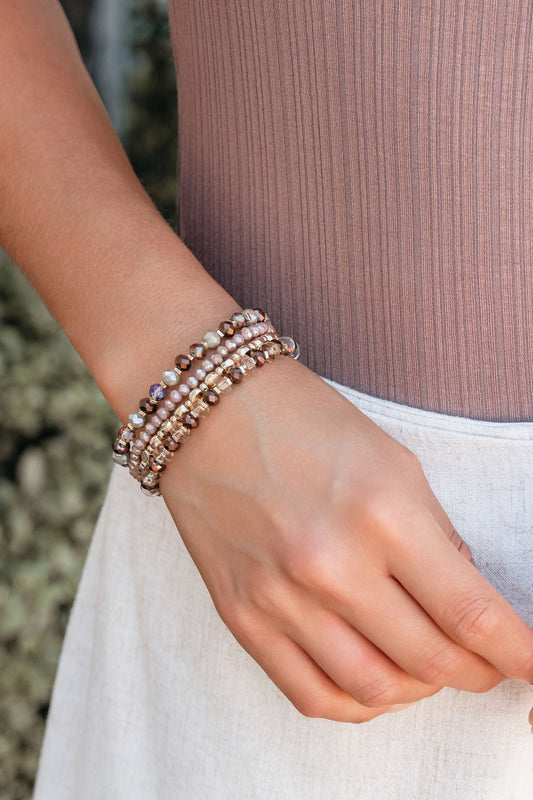 A hand wears the Driftwood Dreams Beaded Layered Bracelets in brown and gold, resting on a light-colored skirt.