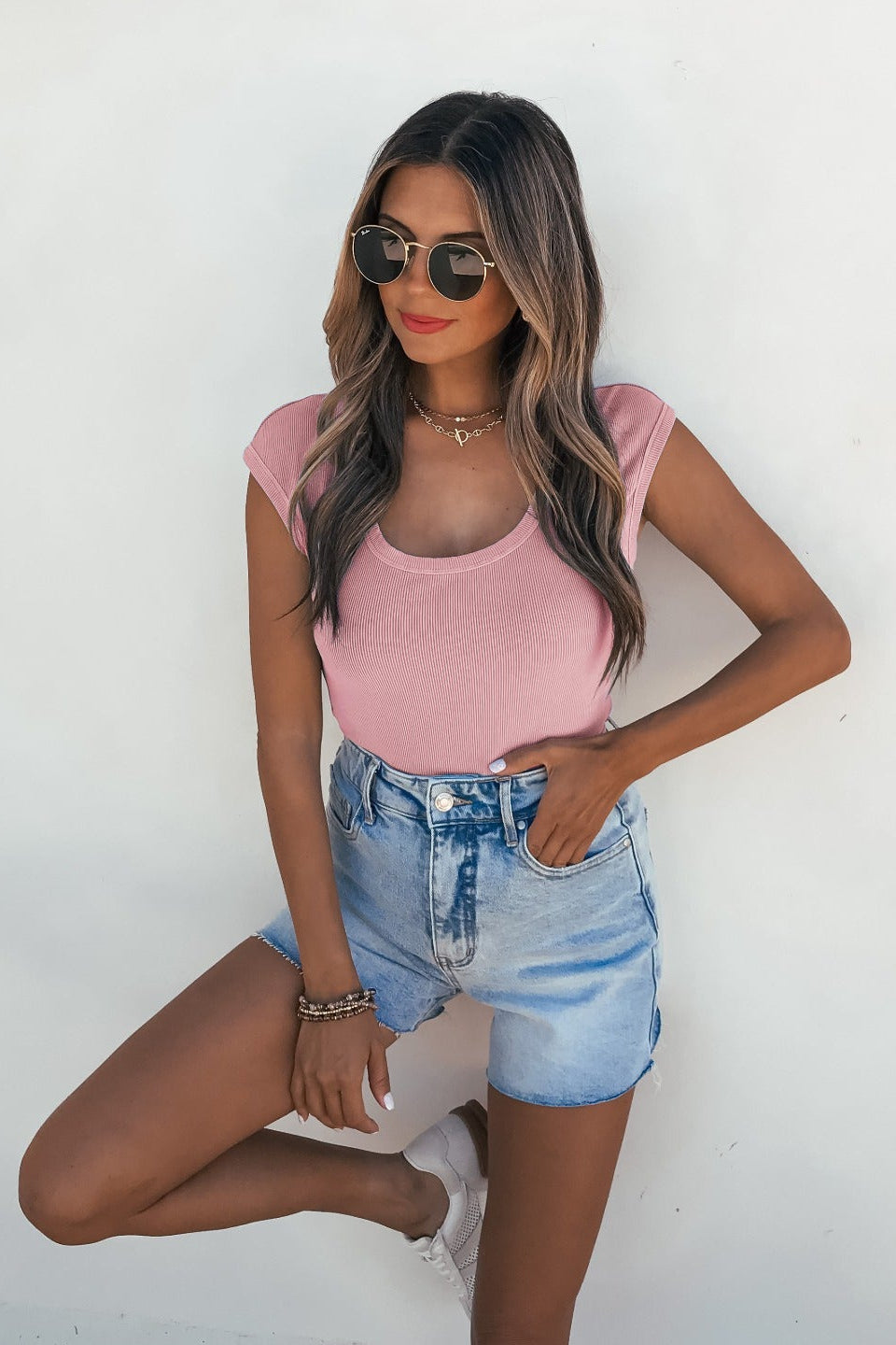 Woman in sunglasses and denim shorts wears a slim Dusty Mauve Scoop Neck Ribbed Top, standing by a white wall with one hand in her pocket.