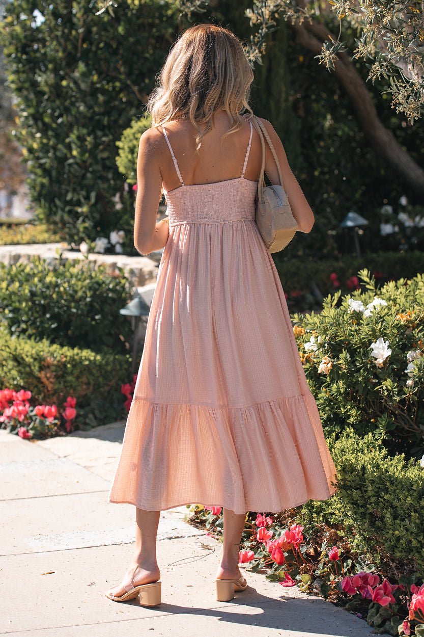 A woman in the Dusty Pink Blossom Tiered Midi Dress and sandals strolls a flower-filled garden path, seen from behind.