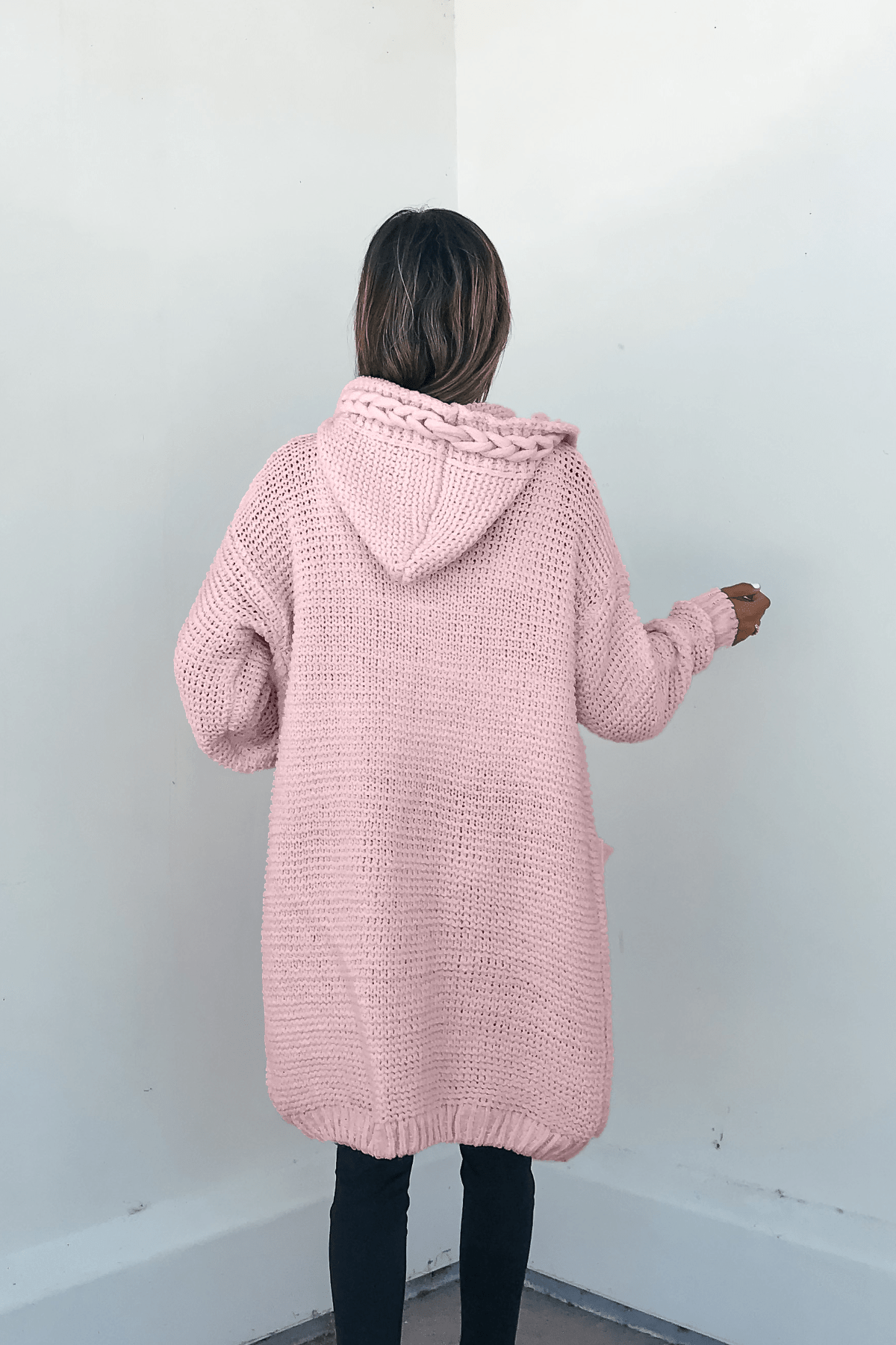 A person in a Dusty Rose Braided Trim Hooded Cardigan faces a light wall, pairing it with dark pants for a cozy knit look.