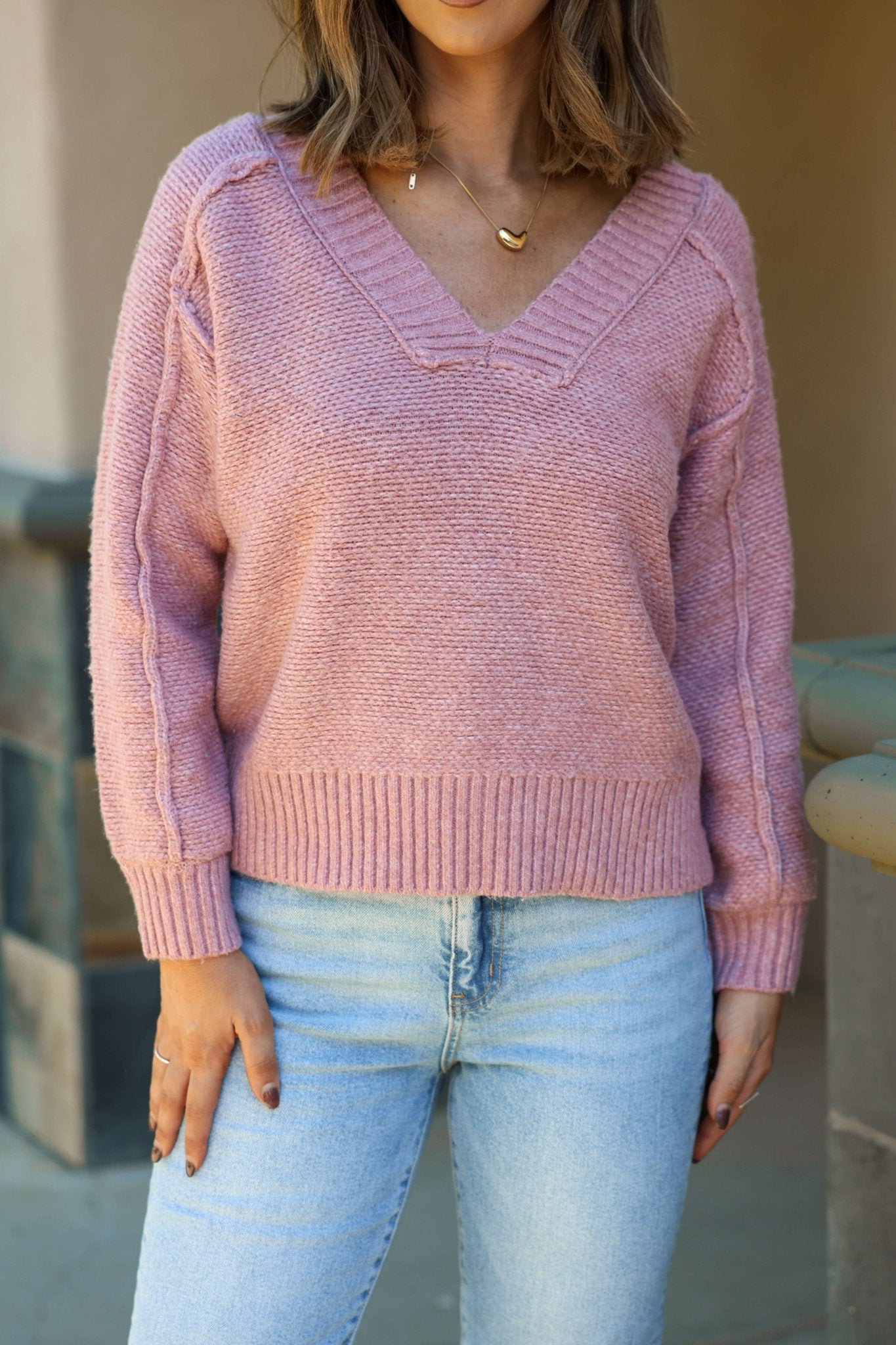 A woman wears a Dusty Rose Seam Detail V Neck Sweater and light blue jeans outdoors, her face partially out of frame.