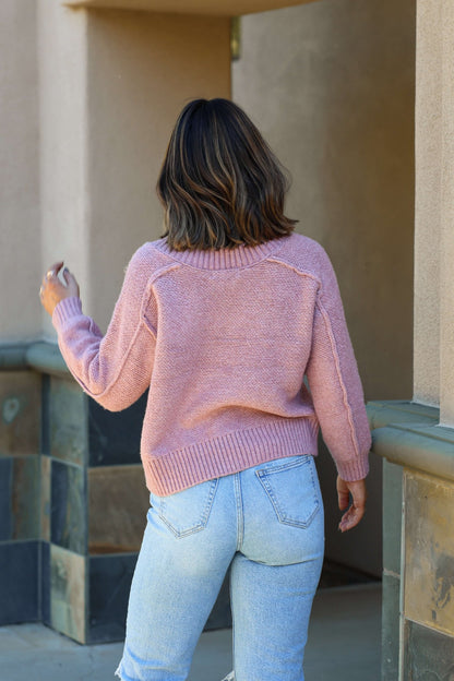 Wearing the Dusty Rose Seam Detail V Neck Sweater and light blue jeans, a person with shoulder-length brown hair walks away.