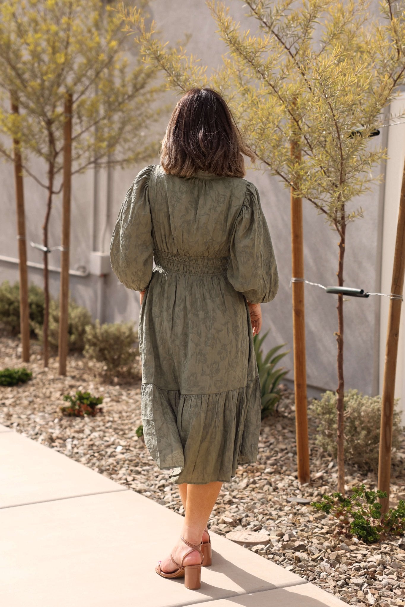 A woman in the Earthy Olive Floral Print Midi Dress walks down a stone path lined with young trees and plants.