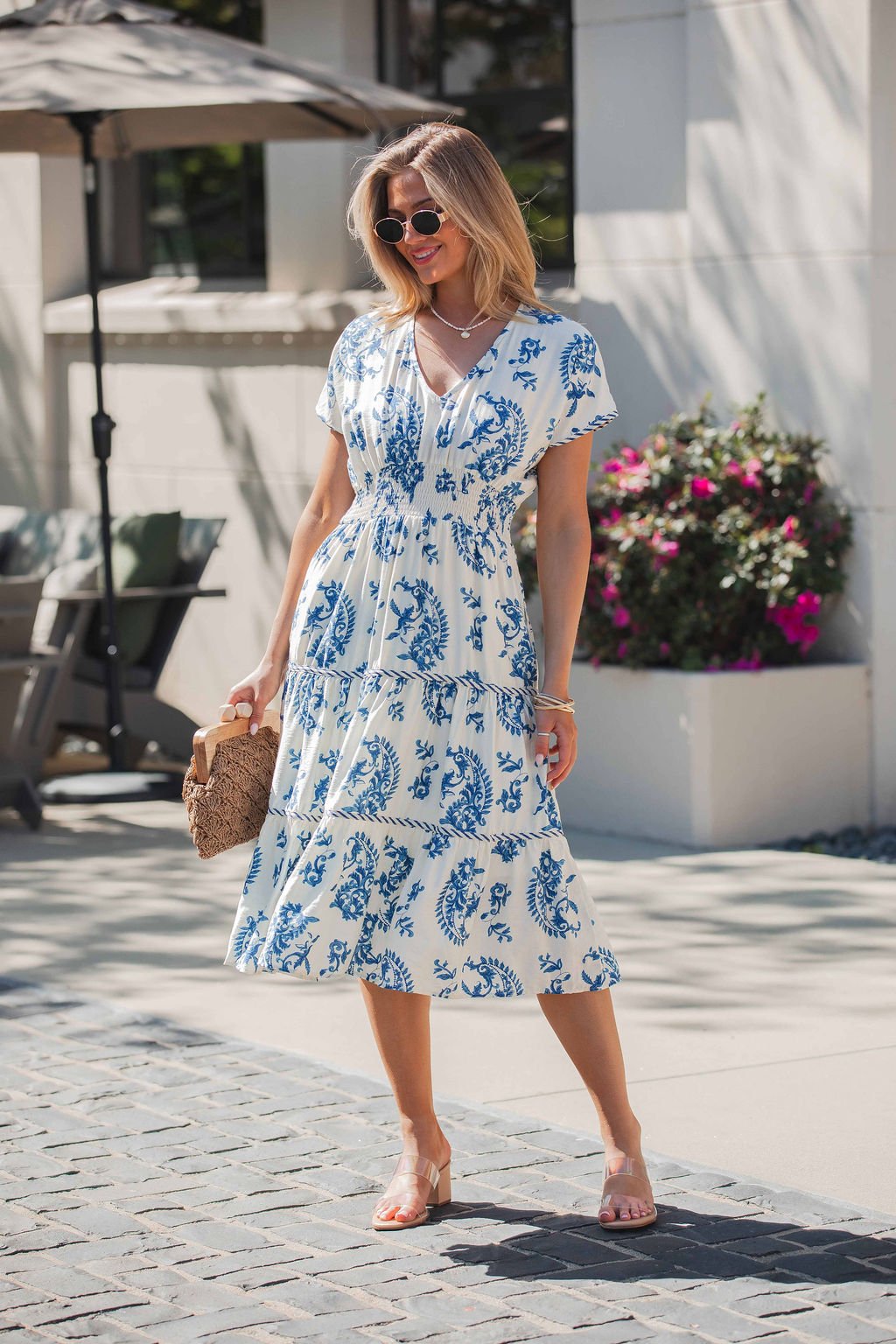 Woman in the Easton Blue Print Smock Tiered Midi Dress, holding a woven bag and wearing sunglasses outdoors on a sunny day.