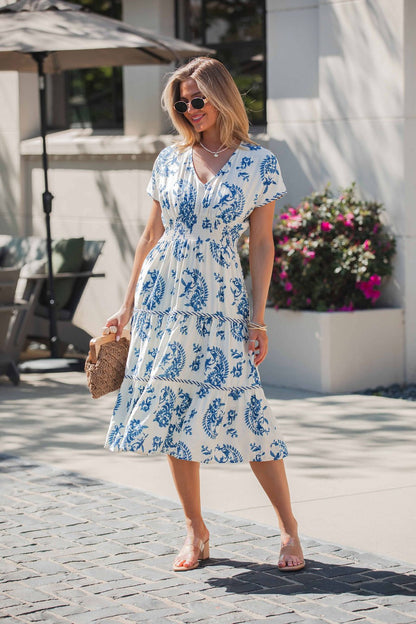 Woman in the Easton Blue Print Smock Tiered Midi Dress, holding a woven bag and wearing sunglasses outdoors on a sunny day.