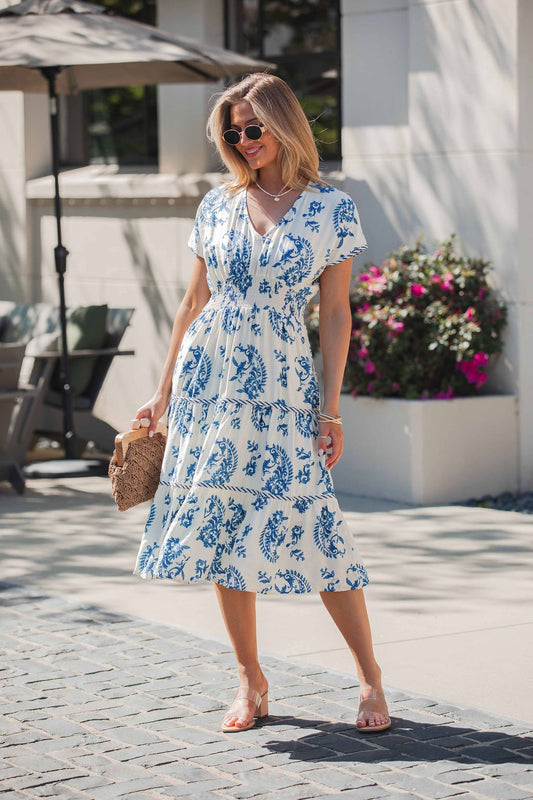 Woman in the Easton Blue Print Smock Tiered Midi Dress, holding a woven bag and wearing sunglasses outdoors on a sunny day.