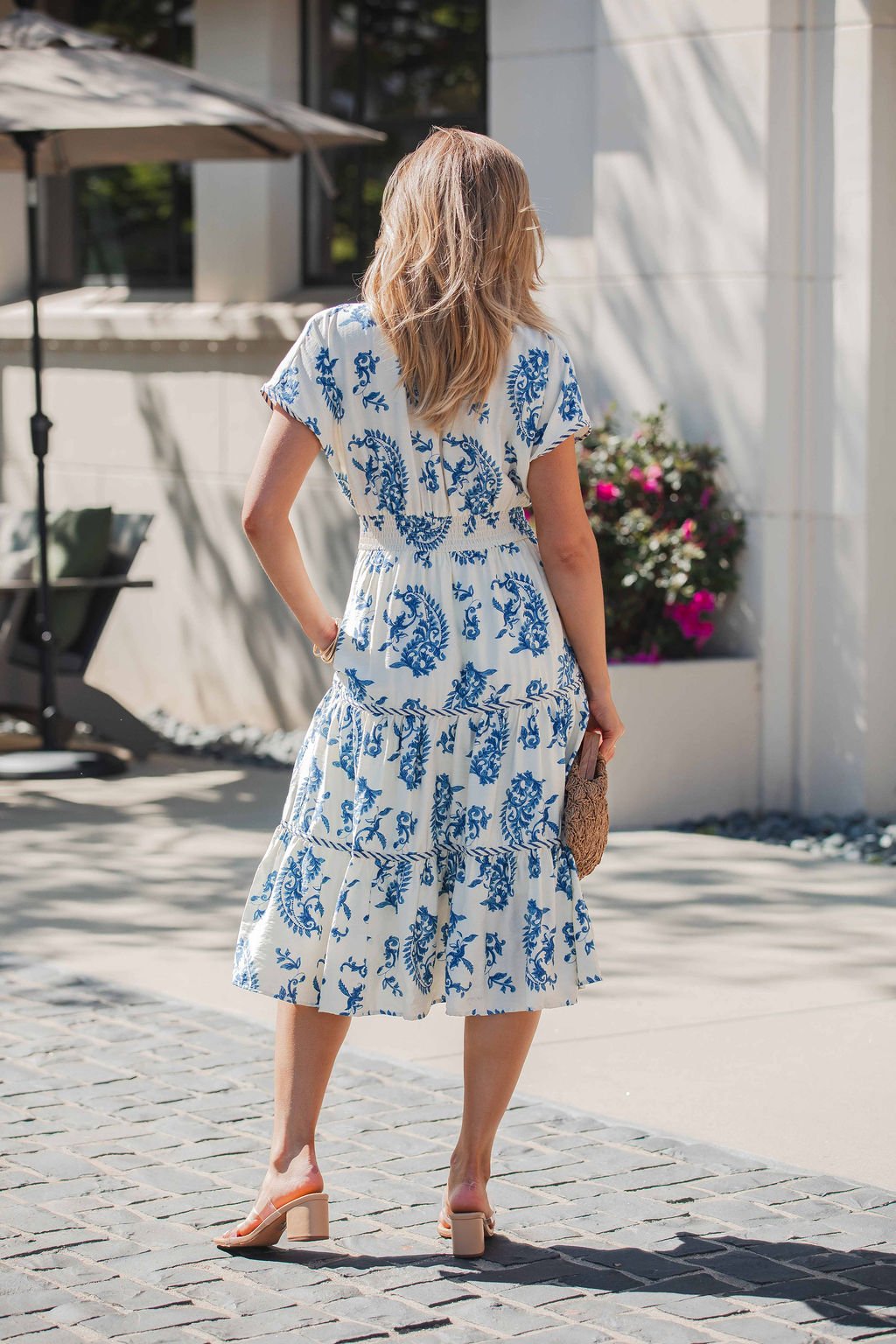 A woman in the Easton Blue Print Smock Tiered Midi Dress stands outdoors, holding a straw clutch.