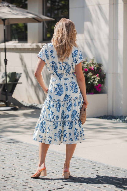 A woman in the Easton Blue Print Smock Tiered Midi Dress stands outdoors, holding a straw clutch.