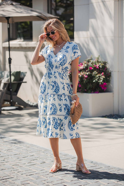 Woman in the Easton Blue Print Smock Tiered Midi Dress, holding a woven clutch, wearing sunglasses and sandals on a sunny day.